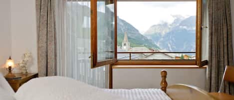 A bedroom with a bed, a nightstand with a lamp, a wooden headboard, and a window with a view of mountains and a church.