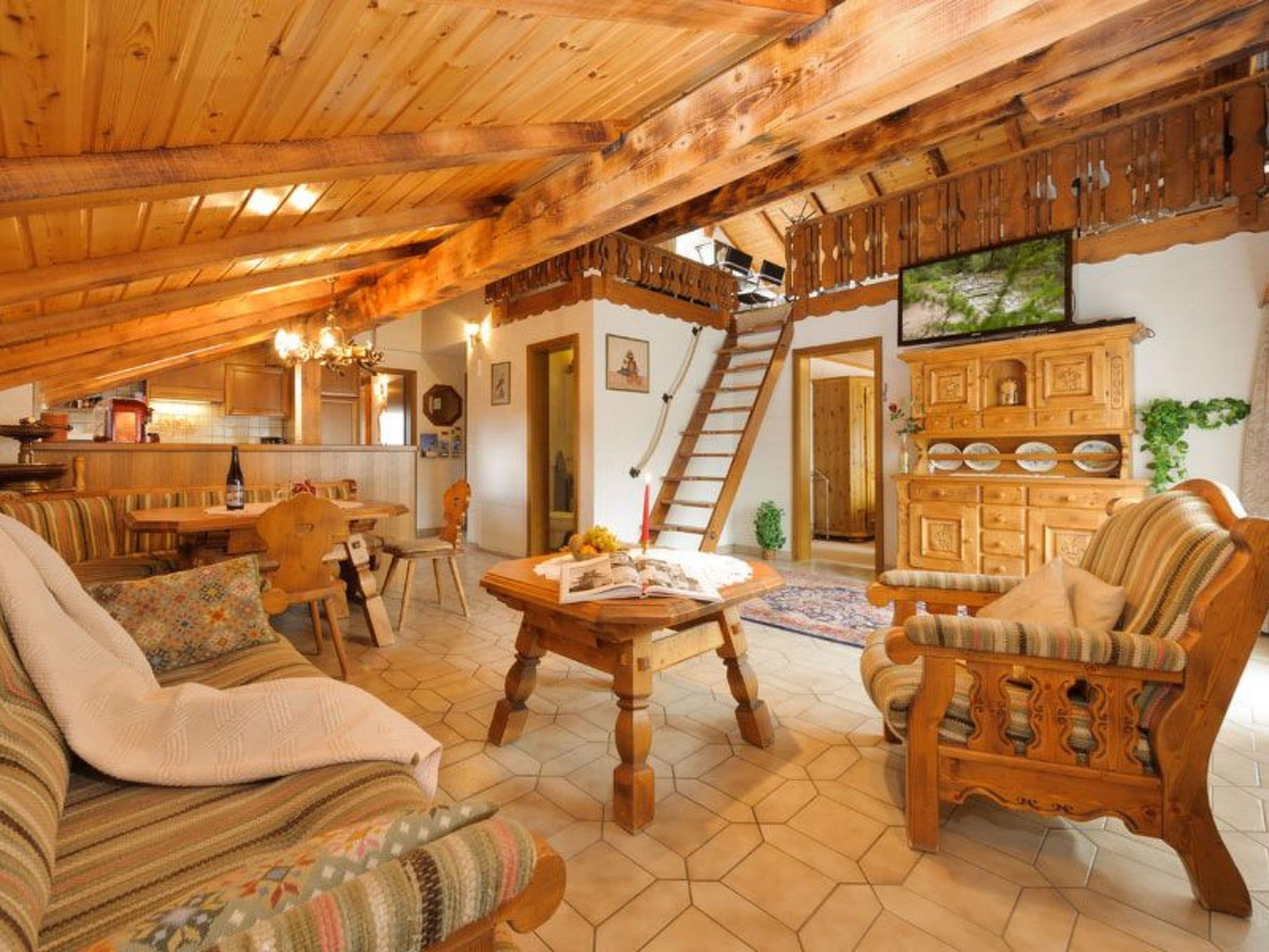 A cozy attic room with a wooden ceiling, a dining area, a sofa, a chair, and a television.