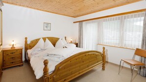 Ein Schlafzimmer mit einem Holzbett, einem Nachttisch, einem Sessel und einem Fenster mit transparenten Vorhängen.