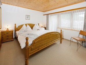 Ein Schlafzimmer mit einem Holzbett, einem Nachttisch, einem Sessel und einem Fenster mit transparenten Vorhängen.