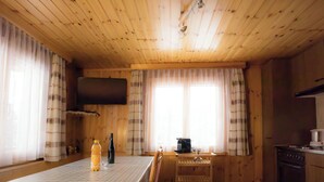 Ein Holzblockbau-Innenraum mit Esstisch, Stühlen, einer Küchenzeile und einem an der Wand montierten Fernseher.