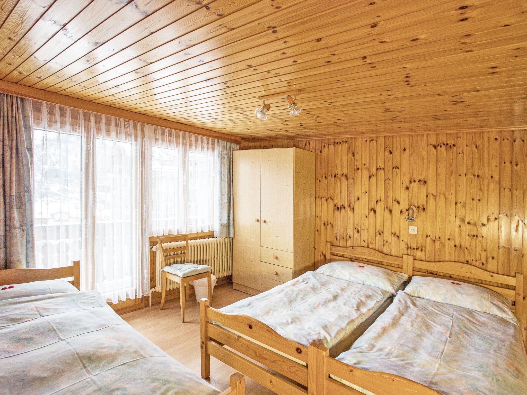 A room with wooden walls and ceiling, two beds, a radiator, a chair, and a window with curtains.