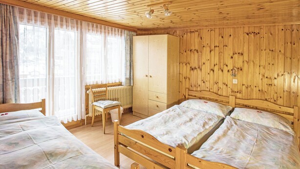 Ein Zimmer einer Holzhütte mit zwei Betten, einem Heizkörper, einem Holzschrank und einem Fenster mit Vorhängen.