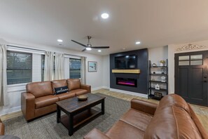 Sala de estar con sofá, mesa de centro, televisor, chimenea, ventilador de techo y estanterías decorativas.
