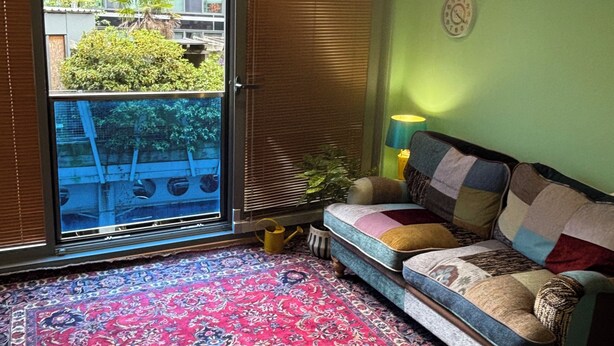 Una habitación con puerta de vidrio corredera que da a un balcón, alfombra estampada, sofá de parches y un reloj de pared.
