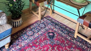 Una habitación con pared verde, un retrato blanco y negro, una mesa de madera con platos verdes, una silla colorida, una planta en maceta y una alfombra estampada.