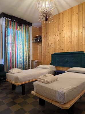 A room with two beds, a chandelier, wooden paneling, and a patterned curtain.