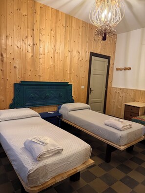 A room with two single beds, a wooden headboard, a chandelier, a door, and a small table.