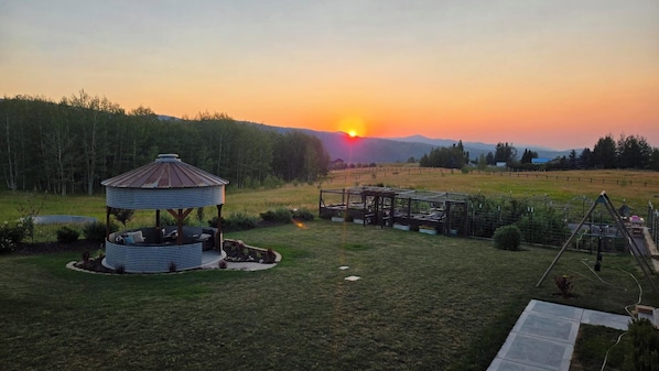 A gazebo with a circular roof and a wooden structure in a grassy field with a sunset in the background.