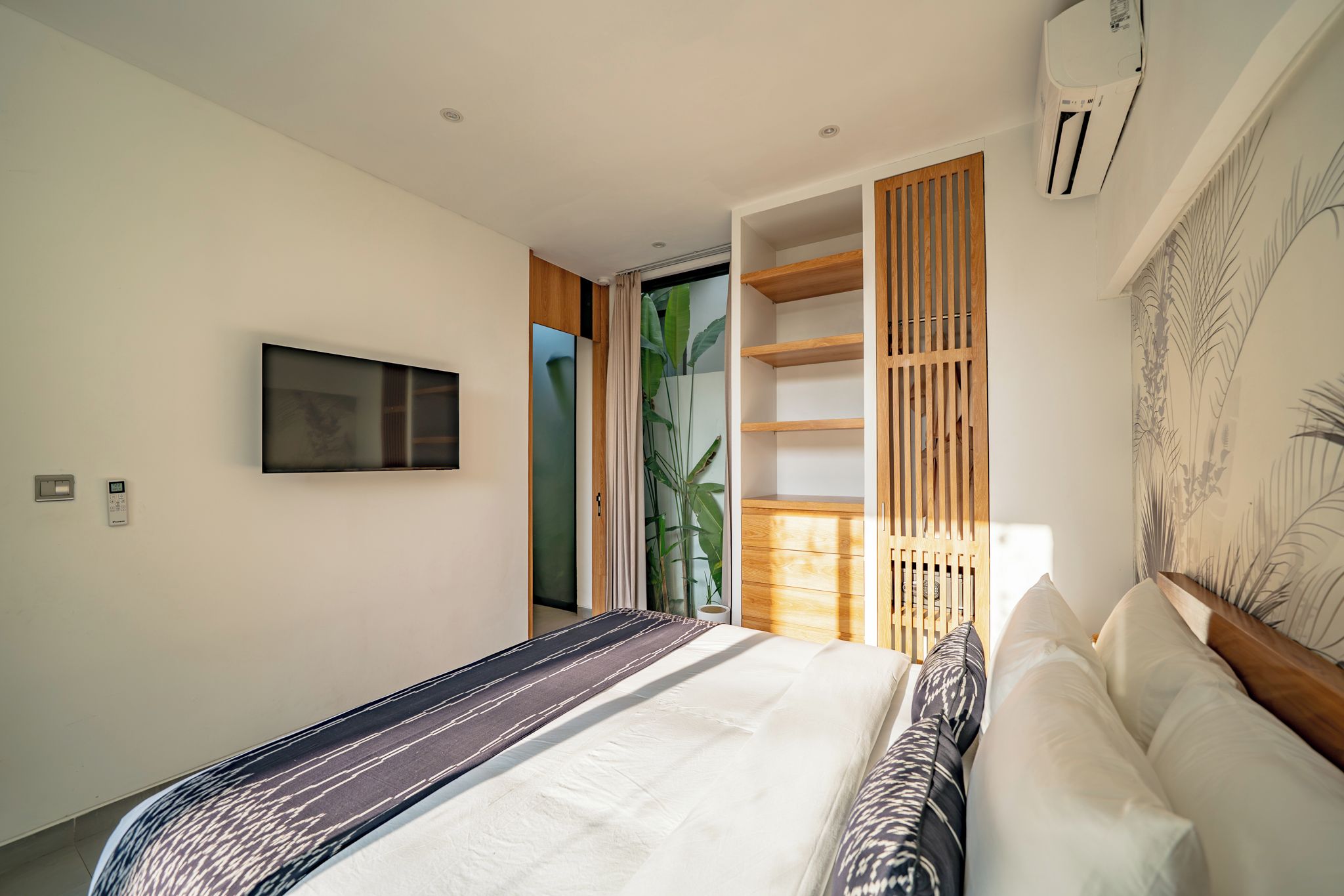 Un dormitorio moderno con cama, televisor en la pared y ventana con vista a follaje.