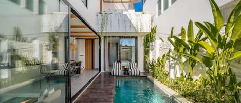 A modern pool area with glass walls, a wooden deck, and a seating area with striped cushions.