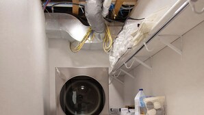 A laundry room with two stacked front-loading washing machines, a drying rack, a basket, and a shelf with cleaning supplies.