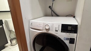 Una lavatrice bianca e compatta con caricamento frontale in una piccola lavanderia con pavimento piastrellato e un tubo dell'acqua a vista.