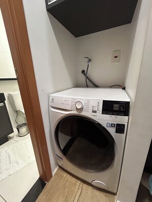 Eine kompakte, weiße Waschmaschine mit Frontlader in einem kleinen Waschraum mit gefliestem Boden und einem sichtbaren Wasserrohr.
