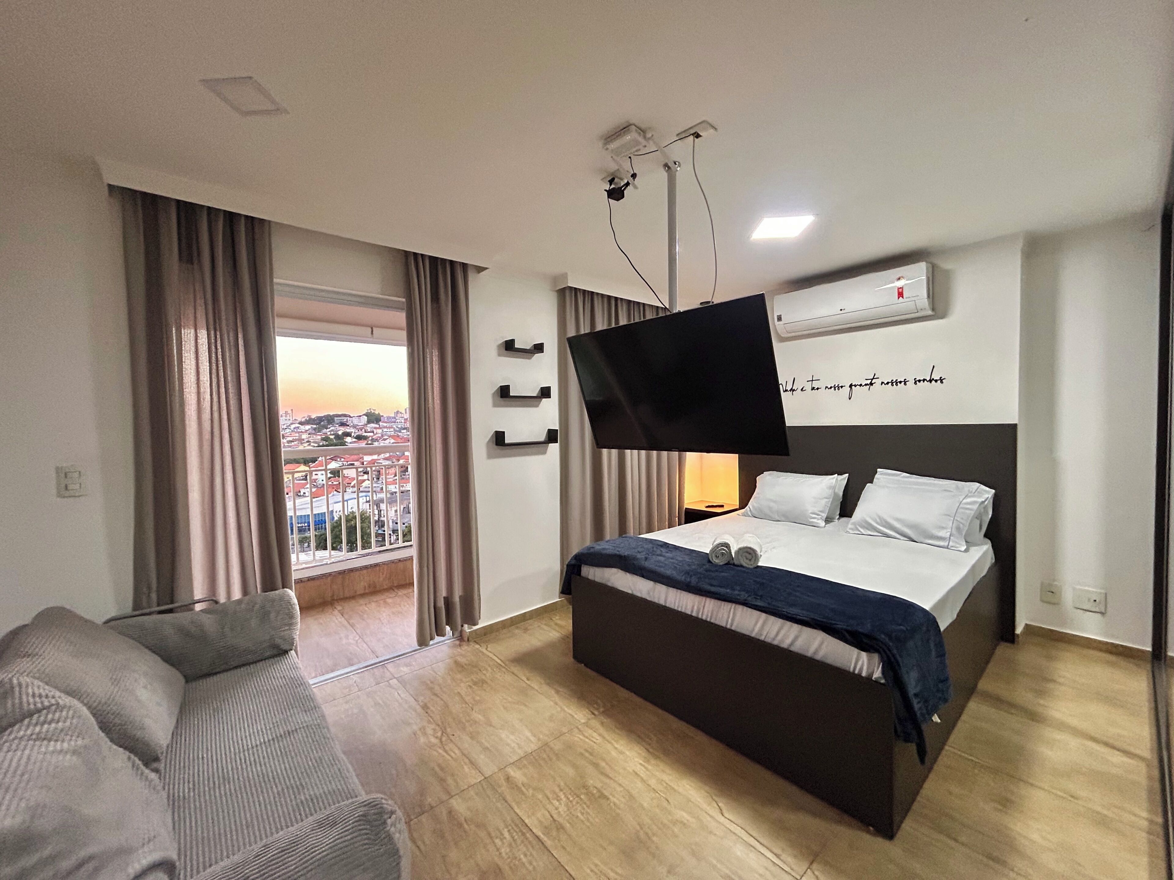 Habitación de hotel con cama, televisor montado en la pared, balcón con cortinas y vista a la ciudad.