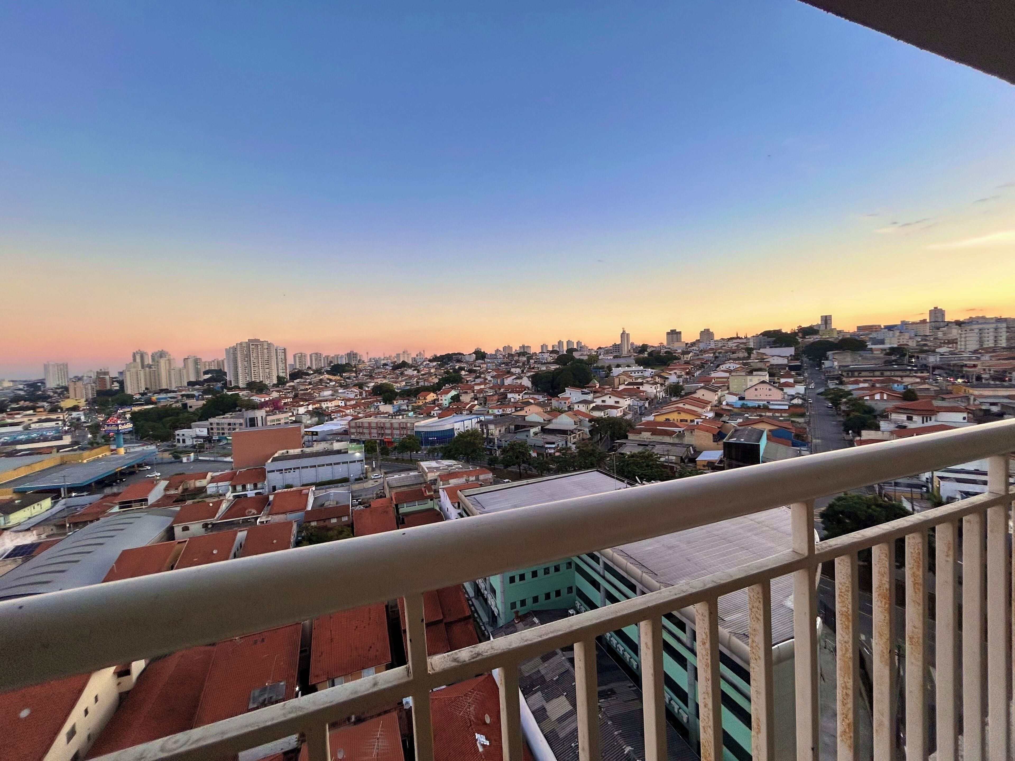 Paesaggio urbano con edifici residenziali e commerciali, cielo sereno e ringhiera di un balcone.