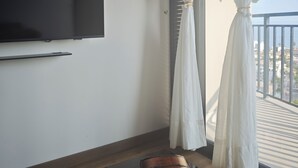 Ein Zimmer mit flachbildfernseher an der Wand, einer Holzbank und einem Balkon mit weißen Vorhängen.