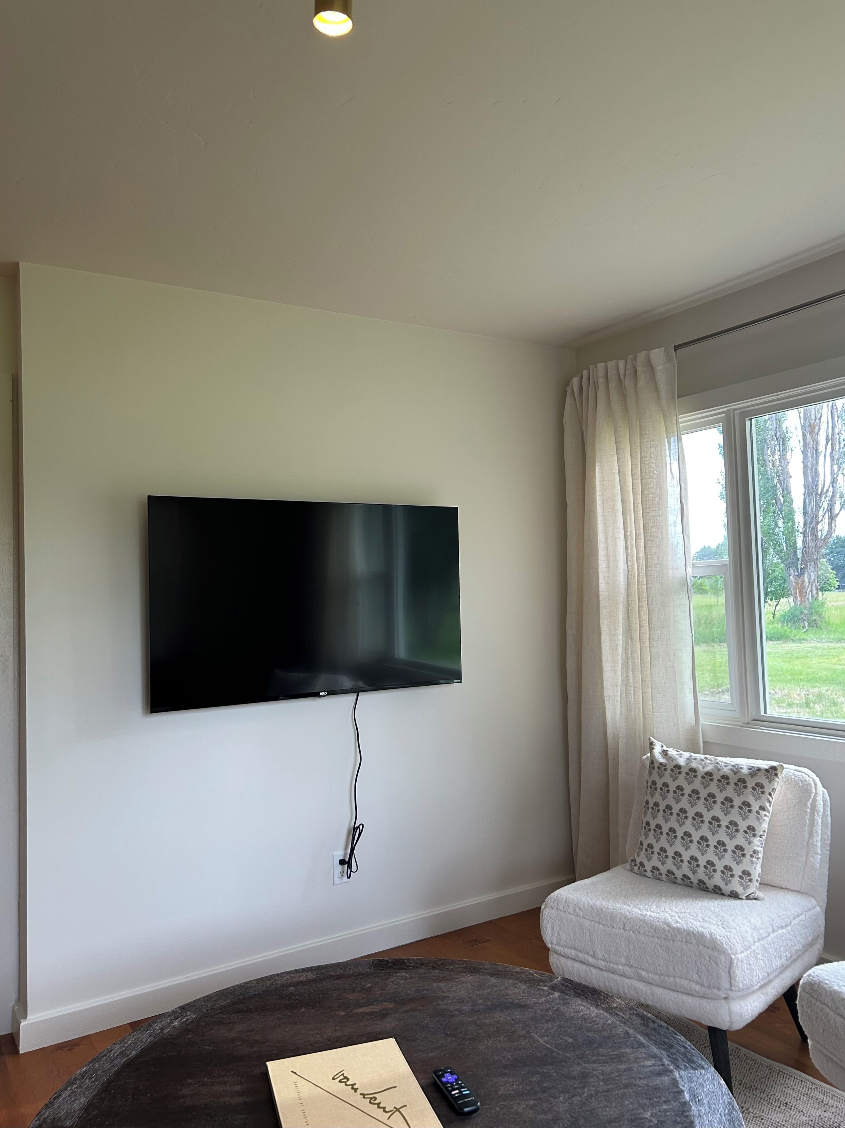A living room with a flat-screen TV mounted on the wall, a round coffee table, a white armchair with a patterned cushion, and a window with curtains.