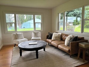 Ein Wohnzimmer mit einem braunen Ledersofa, einem runden Couchtisch und zwei weißen Sesseln. Große Fenster bieten einen Blick ins Freie.