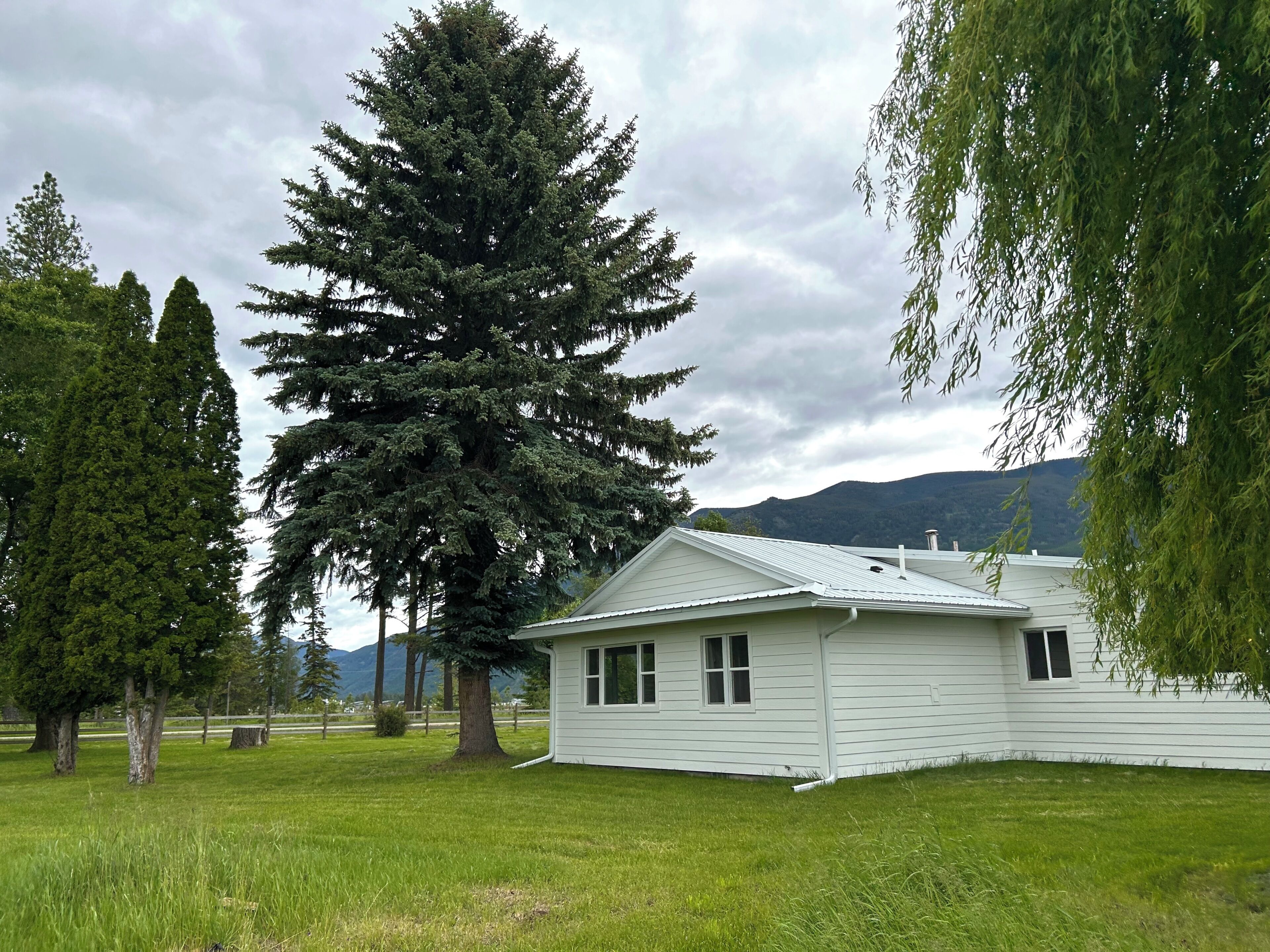 A white single-story house with a sloped roof, surrounded by a grassy yard and tall trees.