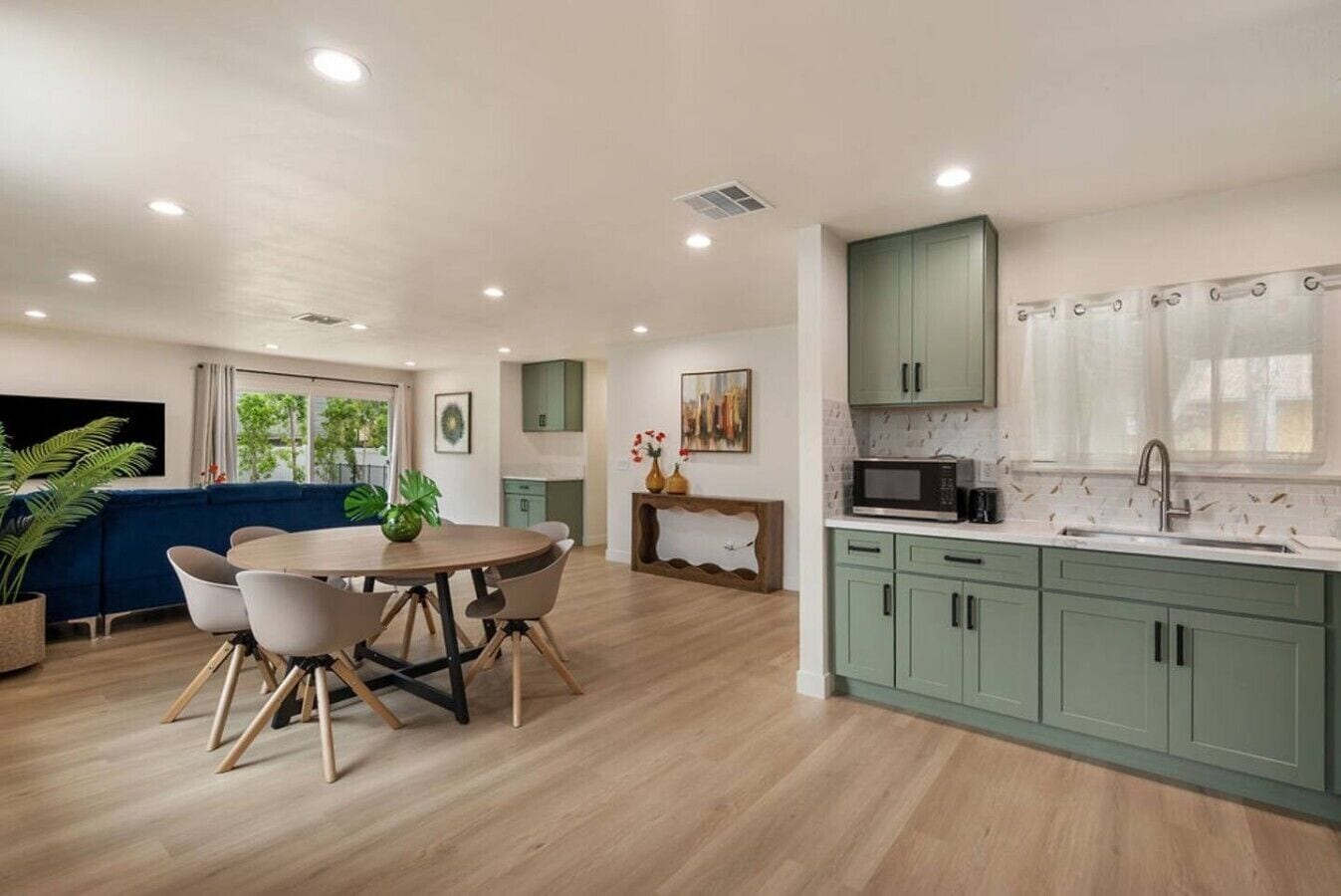 Una cocina moderna con armarios verdes, un comedor con una mesa redonda y sillas, y un amplio ventanal con vistas a la vegetación.