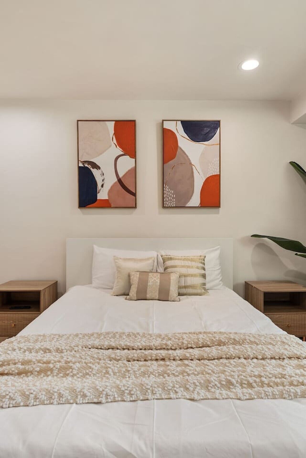 Un dormitorio con cama, almohadas, una colcha, dos cuadros abstractos colgados en la pared y mesitas de noche.