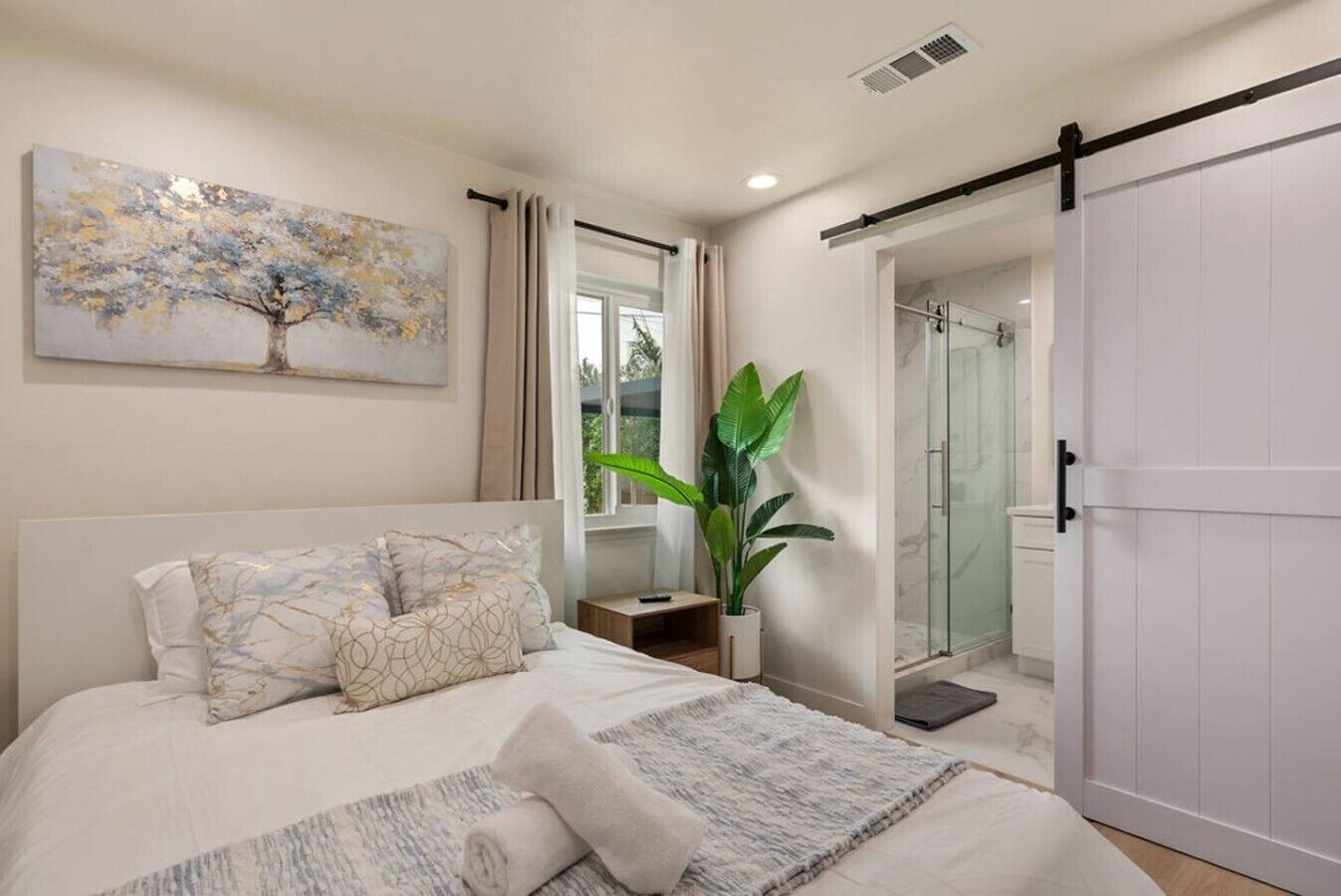 A bedroom with a bed, a painting, a plant, a sliding door, and a shower area.