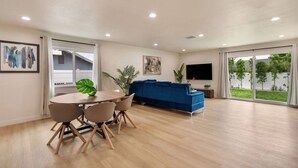 A spacious living area with a dining table, chairs, a blue sofa, a flat-screen TV, and a large window offering a view of the outdoors. - Sunny Nest |Cowboy Pool & Bacyard| Newly Renovated (La Puente)