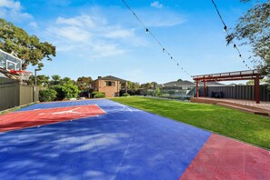 Ein Basketballplatz im Freien mit roter und blauer Spielfläche, umgeben von einem Zaun, Bäumen und einem Gebäude im Hintergrund.
