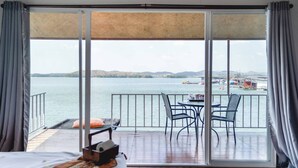 A room with a bed, a table with chairs, and a view of a body of water with boats.