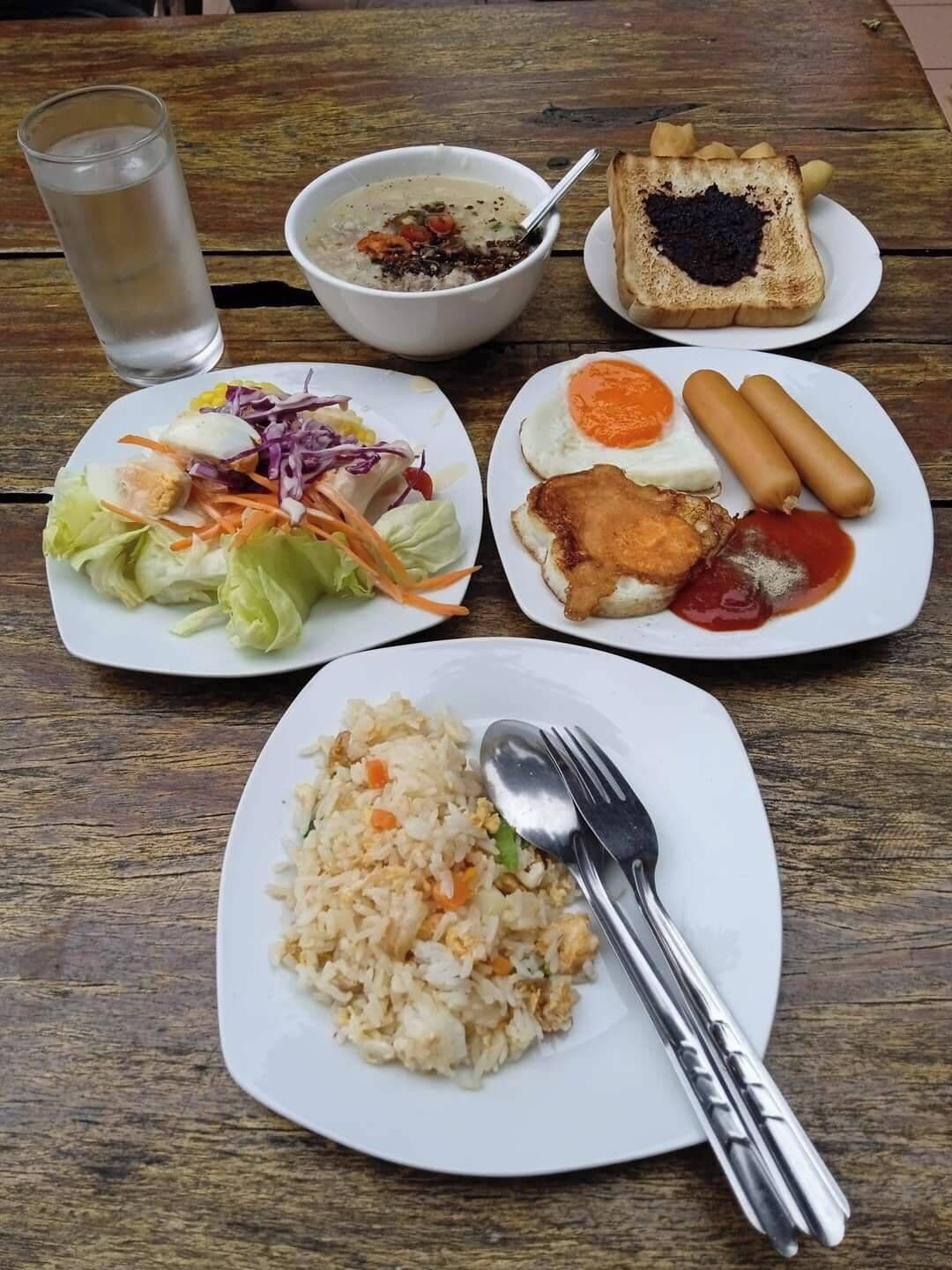 Una mesa con diversos platos, incluyendo un tazón de sopa, arroz frito, una ensalada, un plato con huevos y salchichas, y un vaso de agua.