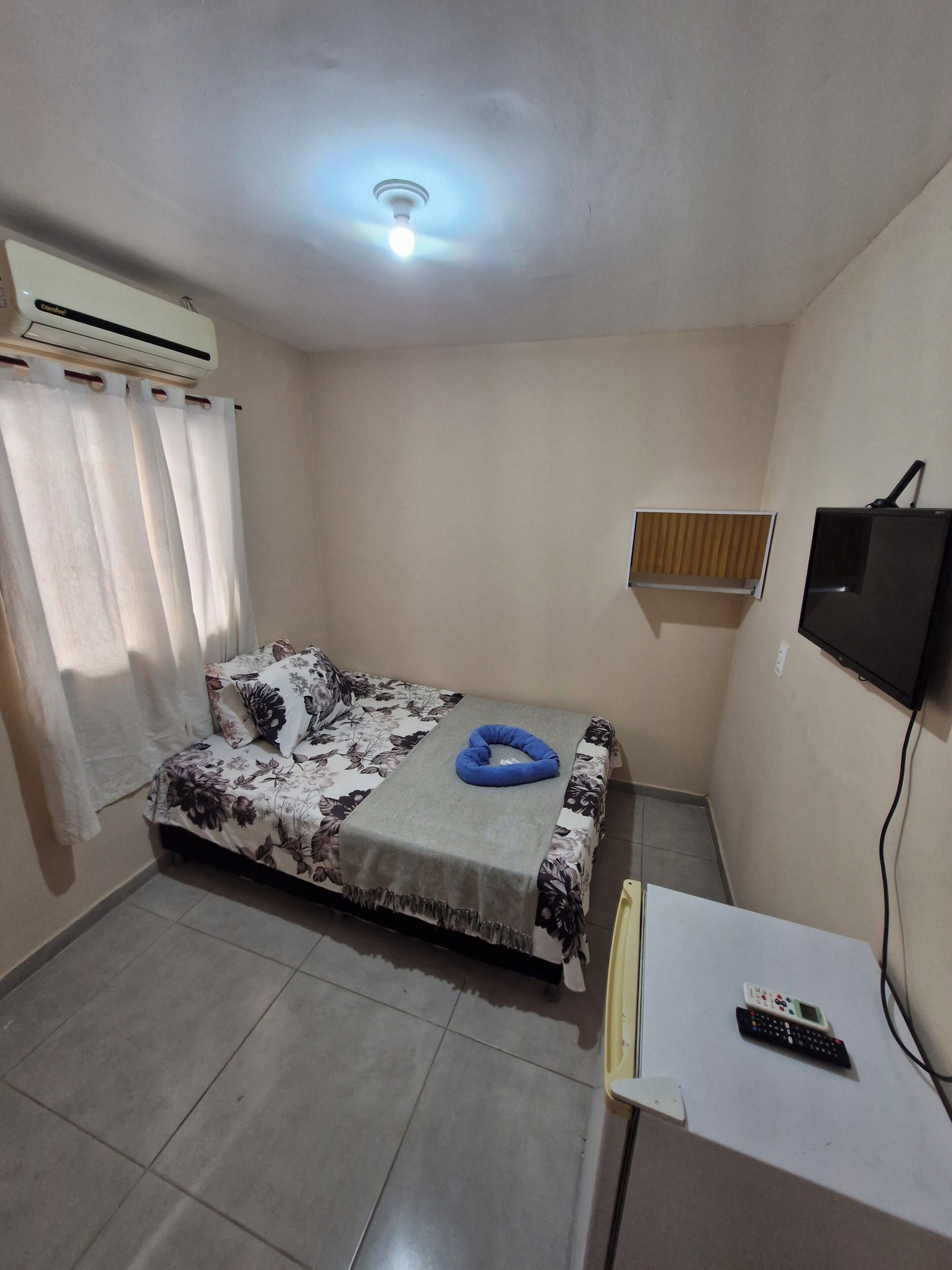 A bedroom with a bed, a TV, a desk with a remote, and an air conditioner.
