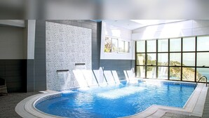 An indoor swimming pool with a tiled wall, a window with a view, and a poolside area. - Hotel Cap Carbon (Béjaïa)