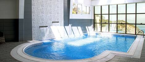 An indoor swimming pool with a tiled wall, a window with a view, and a poolside area.