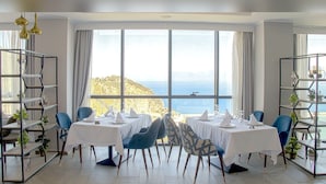 A dining area with tables set for four, large windows with a view of the sea, and metal shelving units with plants. - Hotel Cap Carbon (Béjaïa)