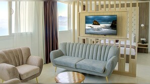 A modern hotel room with a large TV, a sofa, a chair, a small round table, and a bed with a wooden headboard. - Hotel Cap Carbon (Béjaïa)