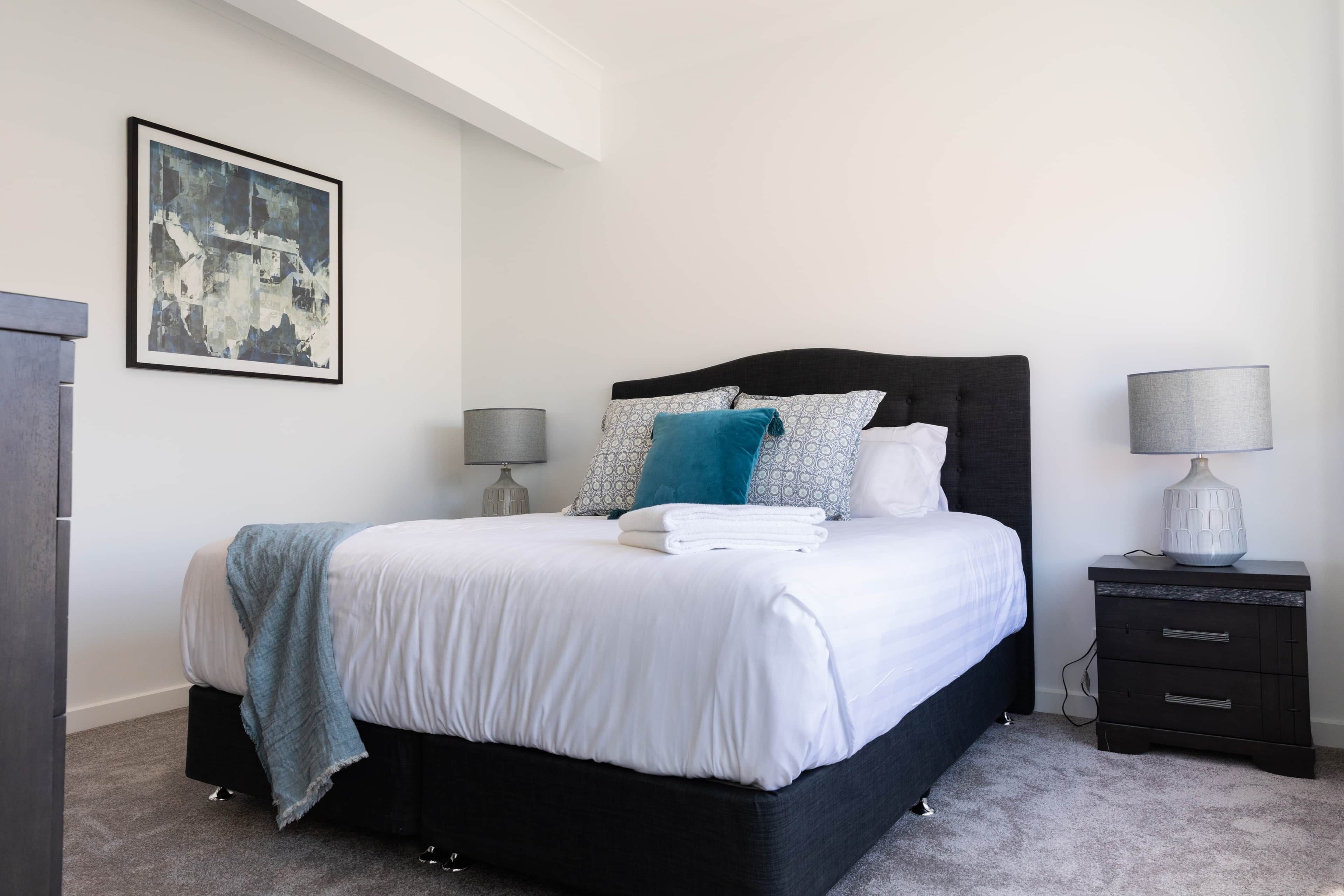 A neatly made bed with a white and blue blanket, a black headboard, and a grey lamp on a bedside table.