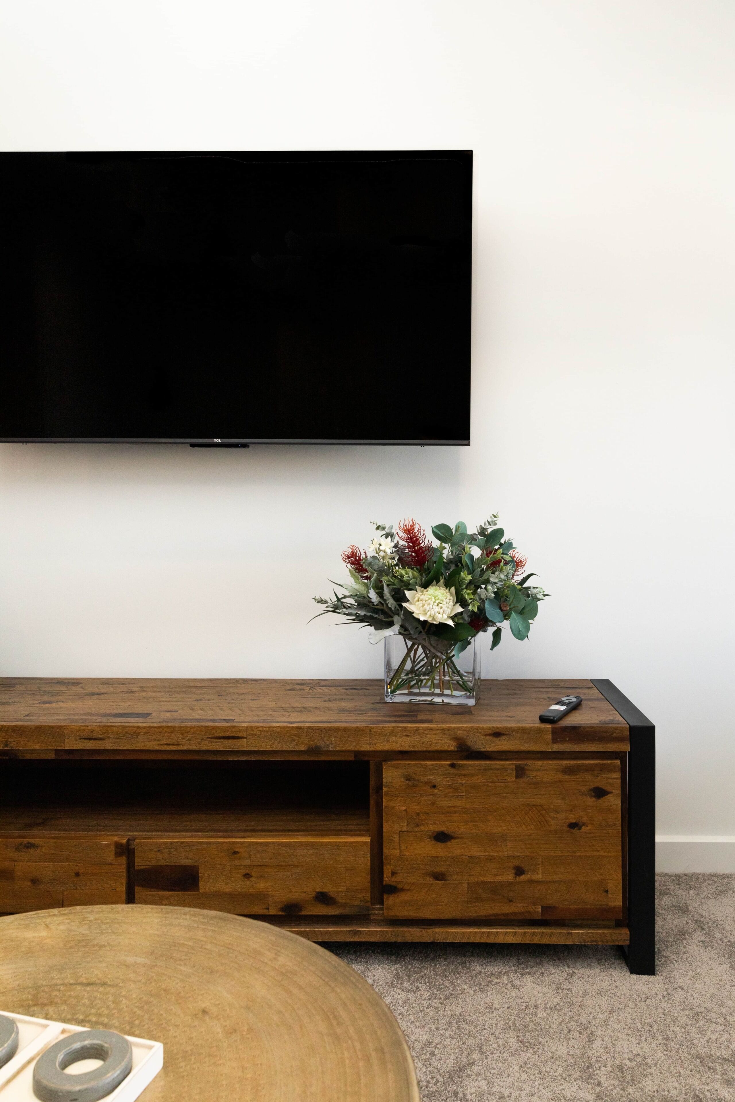 Un moble de roba amb un televisor de pantalla plana muntat a dalt, un vas amb flors al centre, i un comandament a la dreta.