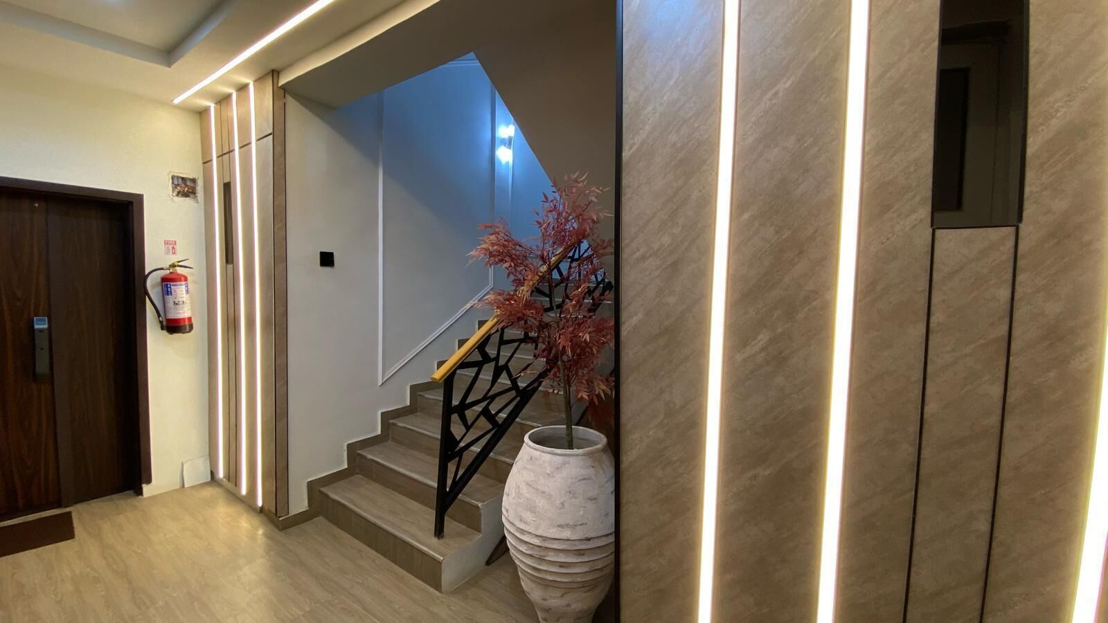 Una escalera moderna con barandilla negra, un jarrón blanco grande con follaje rojo, y un extintor de incendios montado en la pared.