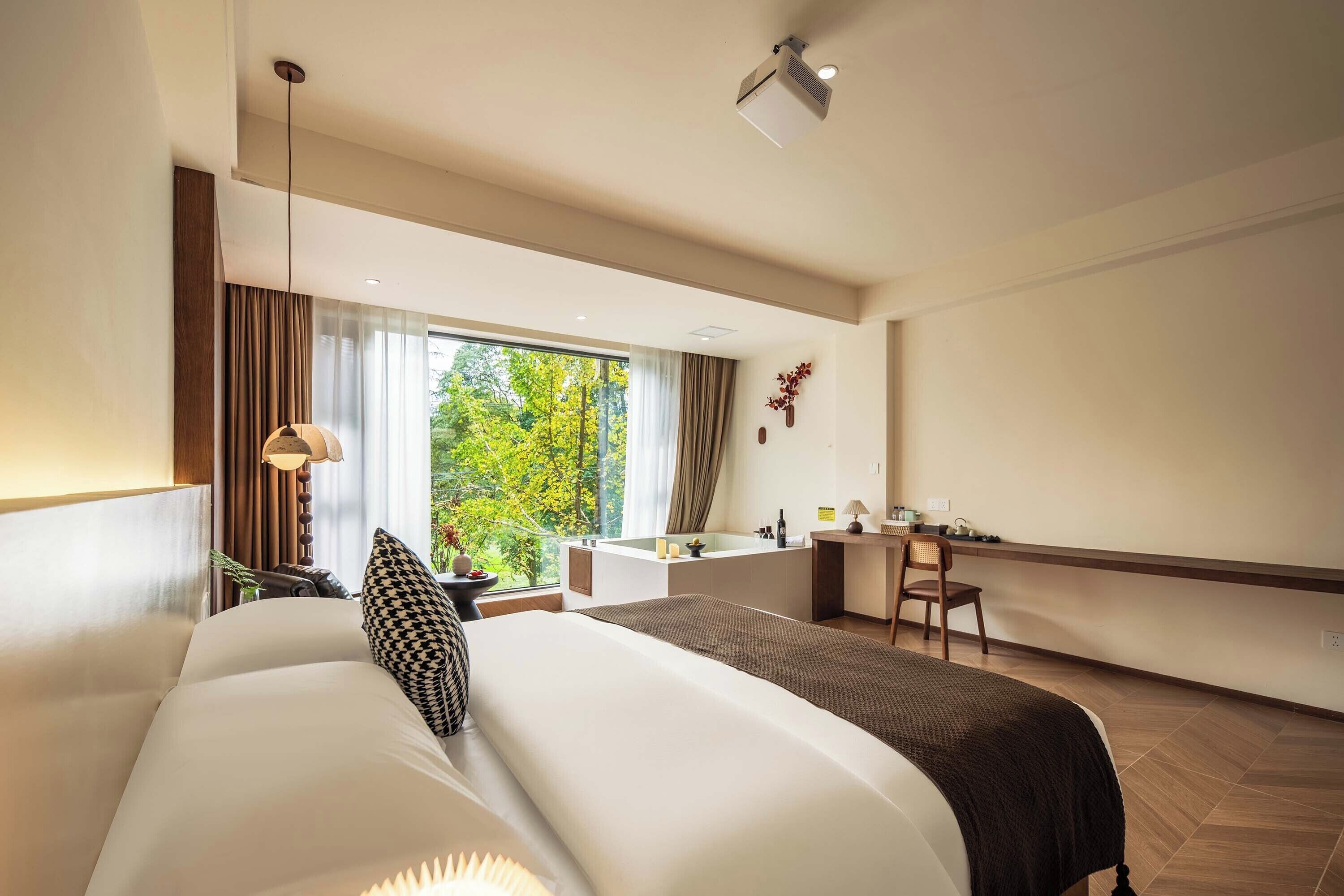 Quarto de hotel com uma cama grande, uma escrivaninha, uma cadeira, uma janela com vista para árvores e um projetor no teto.
