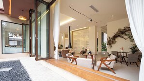 Un interior moderno con entrada por puerta de vidrio, una zona de estar con sofás y sillones, una cocina visible a través del vidrio y una pared decorativa con plantas.