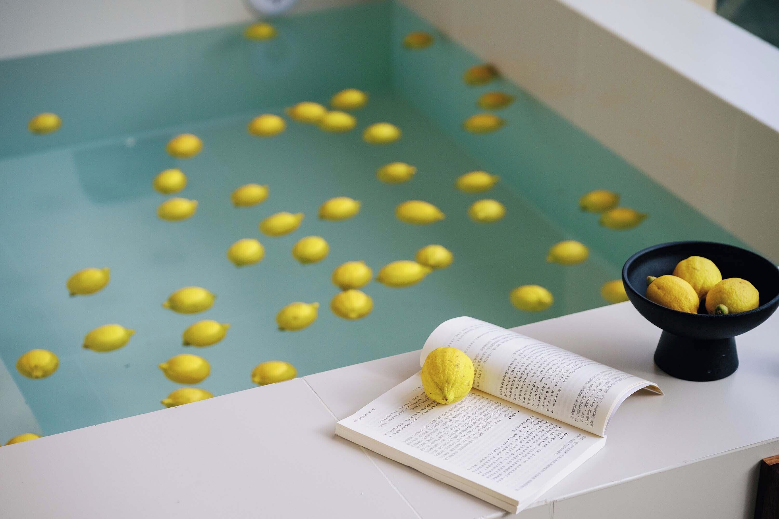 Une baignoire avec des pastilles de bain jaunes, un bol de citrons et un livre ouvert sur une surface blanche.
