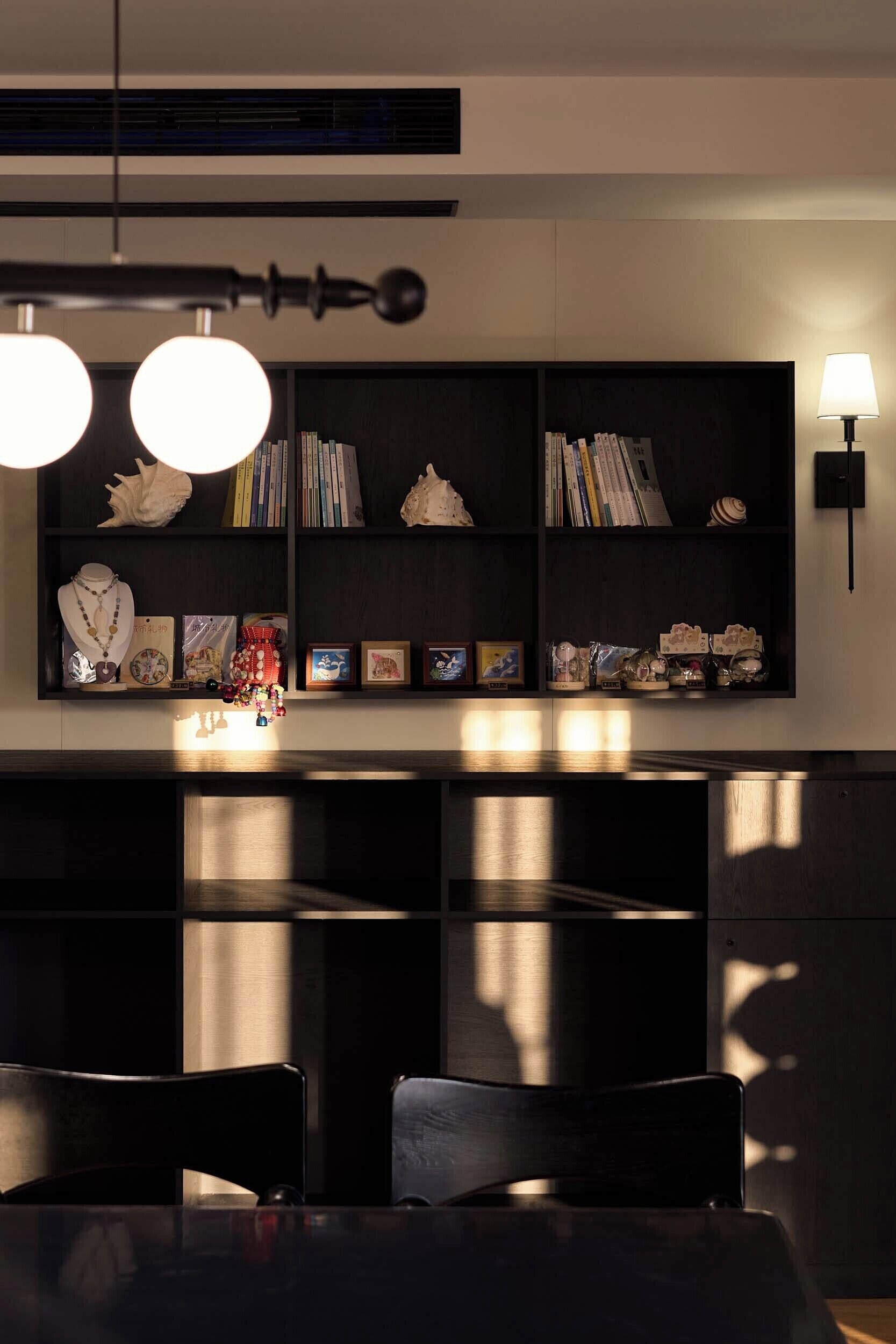 A modern interior with a dark wood dining table, chairs, a bookshelf with decorative items, and a wall-mounted lamp.