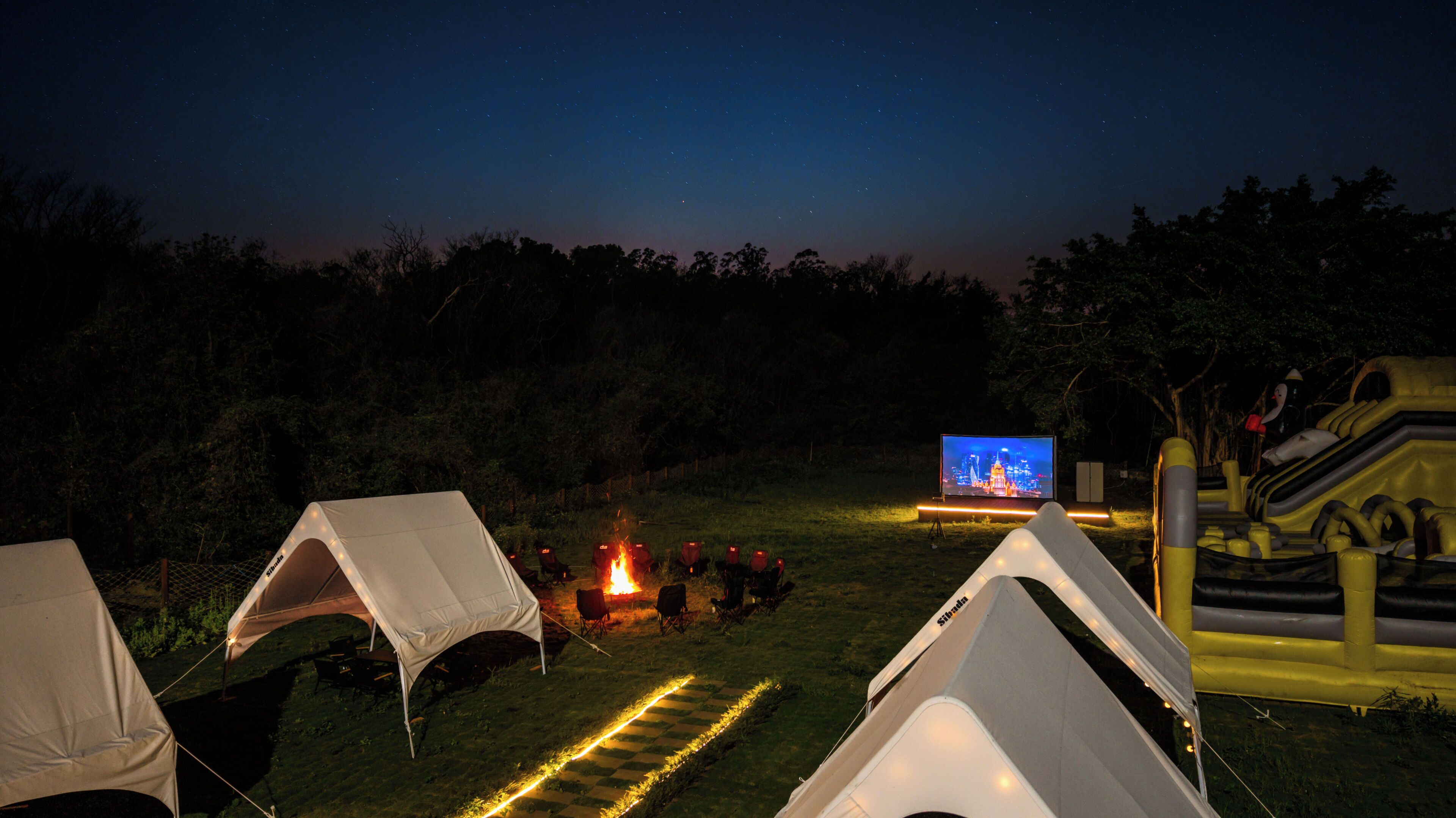 Un terrain de camping de nuit, avec des tentes, un feu de camp et un écran de télévision diffusant une vue sur une ville.