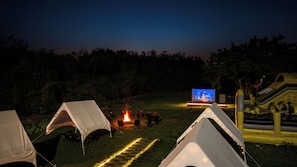 A campsite at night with tents, a campfire, and a TV screen displaying a cityscape.