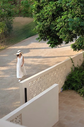 A person in a white dress standing on a balcony overlooking a road with greenery and a tree.
