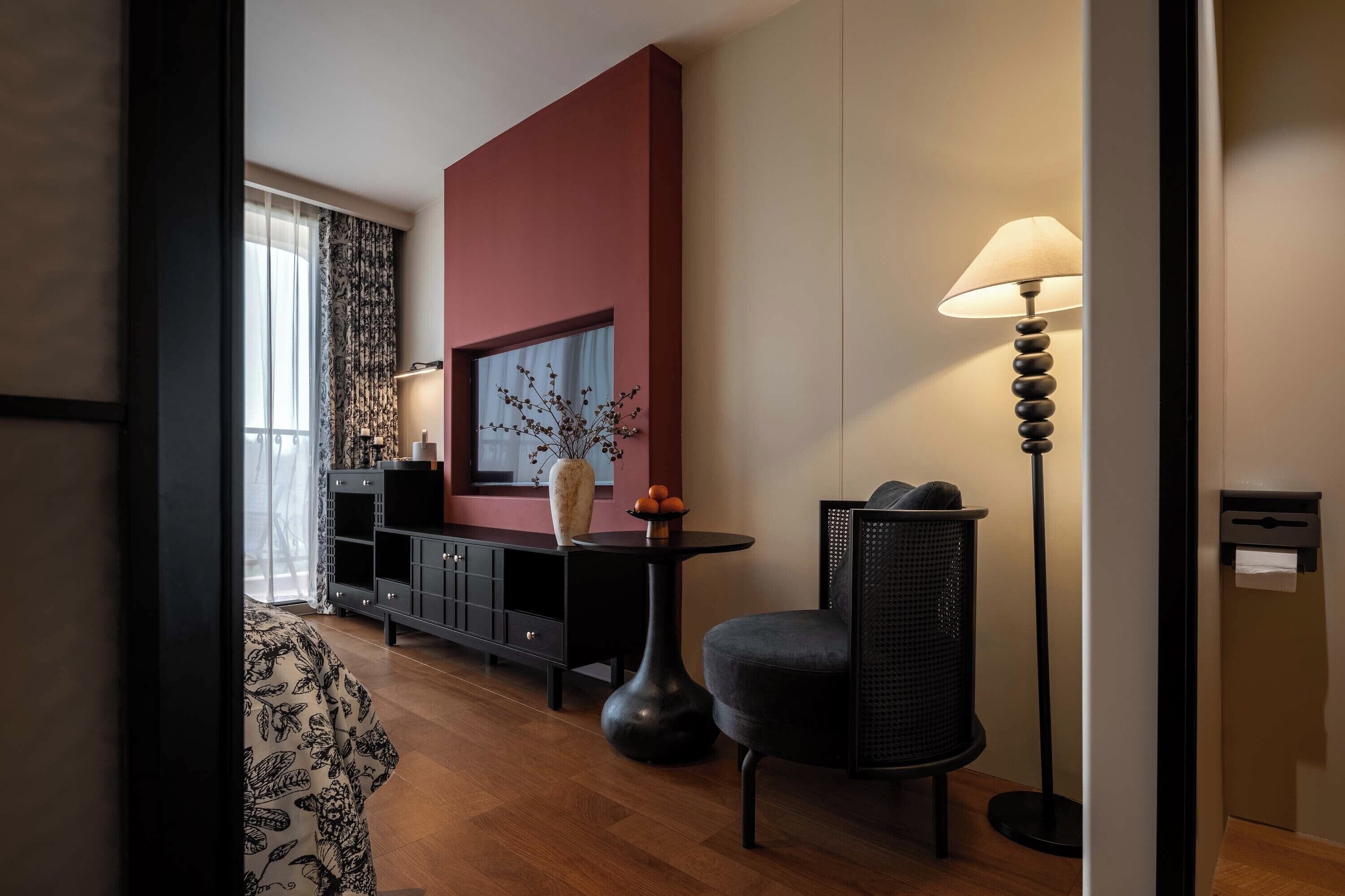 A modern hotel room with a wooden floor, a black sofa, a round table, a floor lamp, a black cabinet, and a large window with curtains.