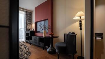 A modern hotel room with a wooden floor, a black sofa, a round table, a floor lamp, a black cabinet, and a large window with curtains.