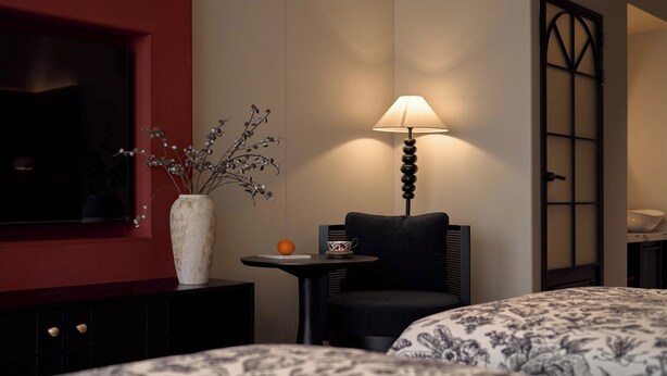 Quarto de hotel com uma cama, uma pequena mesa com abajur, um vaso com galhos e uma televisão em uma parede vermelha.