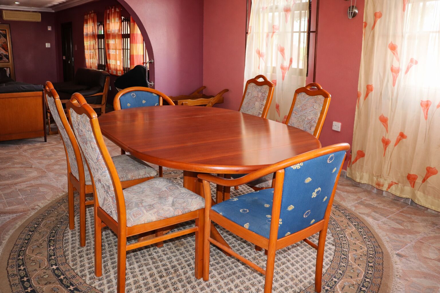 Een eetgedeelte met een houten tafel, zes stoelen, een vloerkleed met dessin en een bank op de achtergrond.